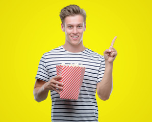 Sticker - Young handsome blond man eating popcorn very happy pointing with hand and finger to the side