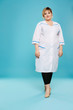 Full length isolated studio portrait of a beautiful chubby woman in a white lab coat. The confident lady posing over the blue background, looking at the camera, smiling. Body positive concept.