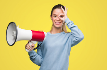 Wall Mural - Beautiful young woman holding megaphone with happy face smiling doing ok sign with hand on eye looking through fingers