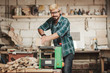 © Screaghin - Attractive young hipster bearded man with eye protectors by profession carpenter builder saws with a circular saw a wooden board on a wooden table in the workshop.