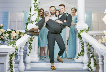 Gay wedding. One groom carrying the other down the steps of the front porch, embellished with flower garlands. Female guests clad in similar blue dresses standing behind, smiles on their faces. 