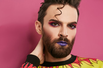 Male makeup look. 3/4 view portrait of a young man rocking beard, quiff, purple lipstick, winged eyeliner, pink eyeshadow. The guy looking at the camera over pink background, his hand on his nape. 