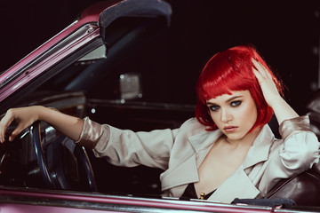 beautiful young woman in red wig and stylish trench coat sitting in car and looking at camera