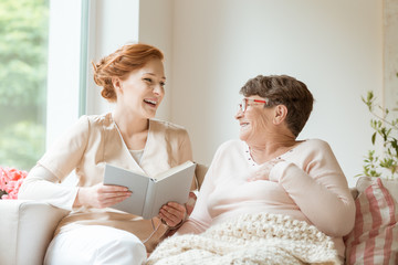 Poster - Happy nurse reading a funny book to her elderly patient in a private nursing home