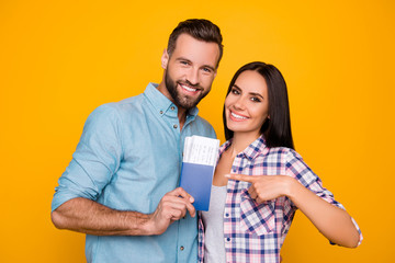 Poster - Portrait of joyful glad couple holding passport with flying tickets in hands pointing with forefinger looking at camera isolated on bright yellow background