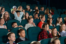 Group Of Stylish Teenagers And Children Expressing Facial Emotions In Movie Theatre. Smiling Spectators Watching Cartoon With Steadfast Eyes And Sitting In Comfortable Blue Chairs.