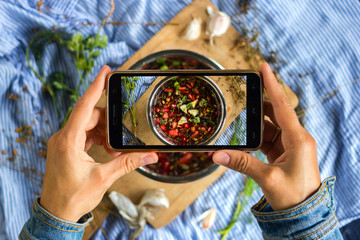 Wall Mural - Woman hands take smartphone food photo of cherry cold chrianteli soup. Phone food photography for social media or blogging in popular and trendy top view style. Raw vegan vegetarian meal concept.