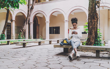 Young Cute Brazilian Undergraduate Girl Is Sitting In The Courtyard Of Her Univercity On The Concrete Bench And Checking Her Schedule And A Hometask Using The Smartphone, Copy Space Area On The Left