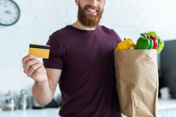 Wall Mural - cropped shot of smiling bearded man holding credit card and grocery bag with vegetables