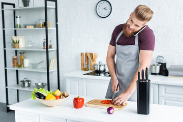 Wall Mural - smiling bearded man in apron talking by smartphone and cutting vegetables in kitchen