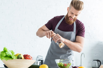Wall Mural - handsome smiling young man in apron spicing vegetable salad