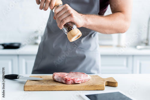 Fotografía cropped shot of man in apron holding pepper mill while cooking beef steak