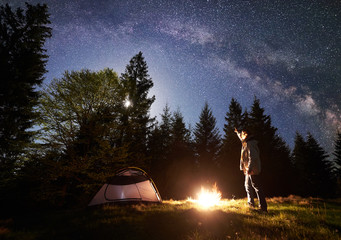 Wall Mural - Camping site in mountain valley at night. Male hiker standing in front of tourist tent at burning campfire, pointing at night blue starry sky with Milky way, full moon. Outdoor activity concept