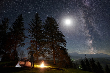 Wall Mural - Camping site in mountain valley. Hiker standing in front of tourist tent at burning campfire near big trees, enjoying night blue starry sky with Milky way and full moon. Tourism and traveling concept