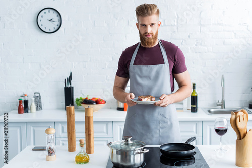 Fotografering handsome bearded young man in apron holding delicious beef steak on plate and lo