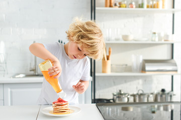 Wall Mural - little boy pouring syrup on pancakes with pieces of strawberry on plate at kitchen