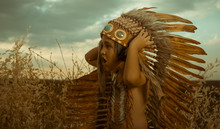 Child Playing At Sunset Dressed As An American Indian, Wearing An Indian Feather Plume And Breastplate. Field Of Wheat And Nature