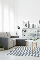 Real photo of a grey couch standing in front of a table that stands on a patterned rug in modern living room interior with shelves with ornaments and posters and a lamp in the bacground