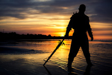 Silhouette Of Assassin With The Sword At The Beach. He Is Posing At The Sea During Beautiful, Orange, Yellow Sunset.
