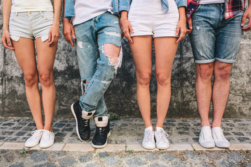 A picture of teens' legs. Three of them are standing straight while man in jeans is holding left leg over right one. Girls wear shorts and one boy as well. They are standing on grey background.