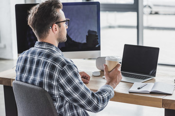 young man in eyeglasses holding paper cup while working with laptop and desktop computer