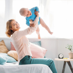 Wall Mural - happy mother raising up laughing little child while sitting on bed at home