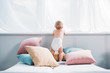 happy little baby in diaper standing on bed with lot of pillows and looking through window