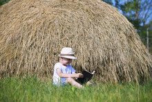 Cute Little Boy Is Reading A Book In A Straw Stack On The Grass In Summer