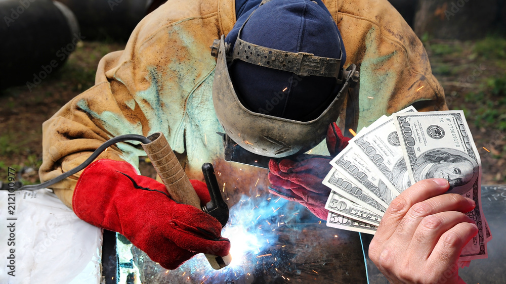 Pipeline Welder Salaries As A Concept / Hand holding US dollar bills in front of an pipeline welder at work with protective equipment - obrazy, fototapety, plakaty 