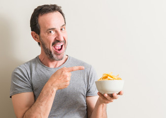 Canvas Print - Senior man eating potato chips very happy pointing with hand and finger to the side