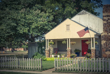 Vintage Modest House With Chimney In Historic Downtown District Of Irving, Texas, USA. Classic Wooden Fence With Well-groomed Landscape, Haning Flower Pots, Big Tree And Proudly American Flag Waving