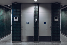 Dark Public Toilet Entrances For Men And For Women In A Modern Airport Terminal Building Or Railway Station Depot, With Greenish Ceramic Tiles On The Wall: Two Dors And Two More Entrances