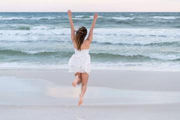 Young happy woman in white dress on beach pink sunset in Florida panhandle with wind, ocean waves, jumping running mid-air to water