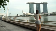 Woman Walks Along The River Embankment And Takes Pictures Of The Marina Bay Sands Hotel In Singapore