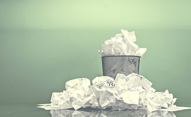 Waste paper bin overflowing with paper against a green wall.