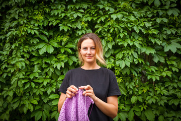 A young woman in a black T-shirt and jeans shorts stands knitting needles and an untied sweater made from natural lilac wool threads against the background of a green wall of grapes