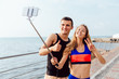 Happy attractive couple in sportswear showing a thumb up and peace sign while taking a selfie on mobile phone, after workout outdoors, near the sea.