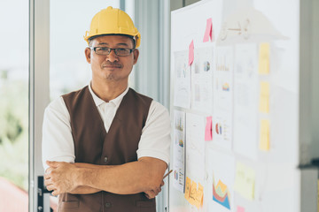 old architect wearing a protective helmet standing in office building with his present white board