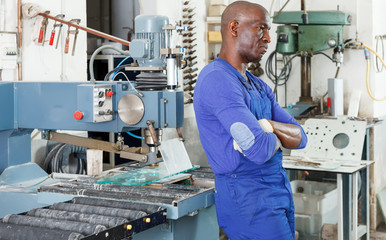 Confident African American workman in blue overalls standing next to glass processing machine