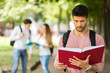 © Minerva Studio - Male student reading a book in a college courtyard with his friends in tha background