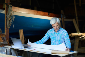 Wall Mural - Experienced shipbuilding engineer looking at sketch on unfolded blueprint by his workplace