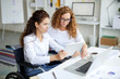 © pressmaster - Young woman in wheelchair and her colleague discussing online data by desk in office