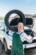 © RomanR - Woman mechanic with tyre on shoulder and broken car