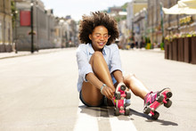 Love Roller-skating. Cute Curly-haired Girl Sitting On The Asphalt In The Middle Of The Street And Putting On Roller Skates