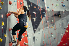 Businesswoman Having Active Recteation Time After Hard Work In Office, Brunette, Dressed In Black Sportweat, Climbing Up On High Rock Artificial Wall.