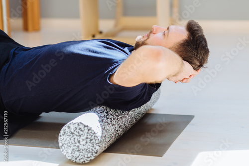 Handsome man performing back exercise on a foam roller being assisted at pilates studio. Patient doing fascia exercise with foam roller on back. Rehabilitation after trauma concept.