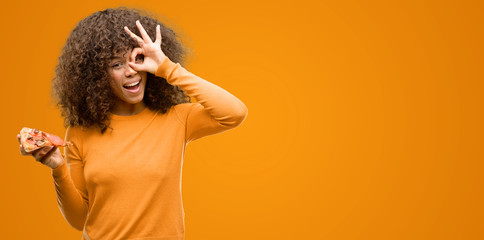 Poster - African american woman with a pizza slice with happy face smiling doing ok sign with hand on eye looking through fingers