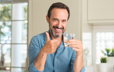 Middle age man drinking a glass of water happy with big smile doing ok sign, thumb up with fingers, excellent sign