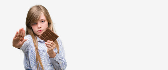 Sticker - Young blonde child holding chocolate bar with open hand doing stop sign with serious and confident expression, defense gesture