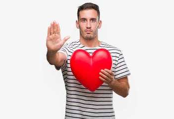 Canvas Print - Handsome young man in love holding red heart with open hand doing stop sign with serious and confident expression, defense gesture